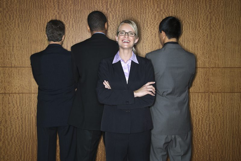 Caucasian businesswoman stands smiling as businessmen stand with their backs turned. Horizontal shot.